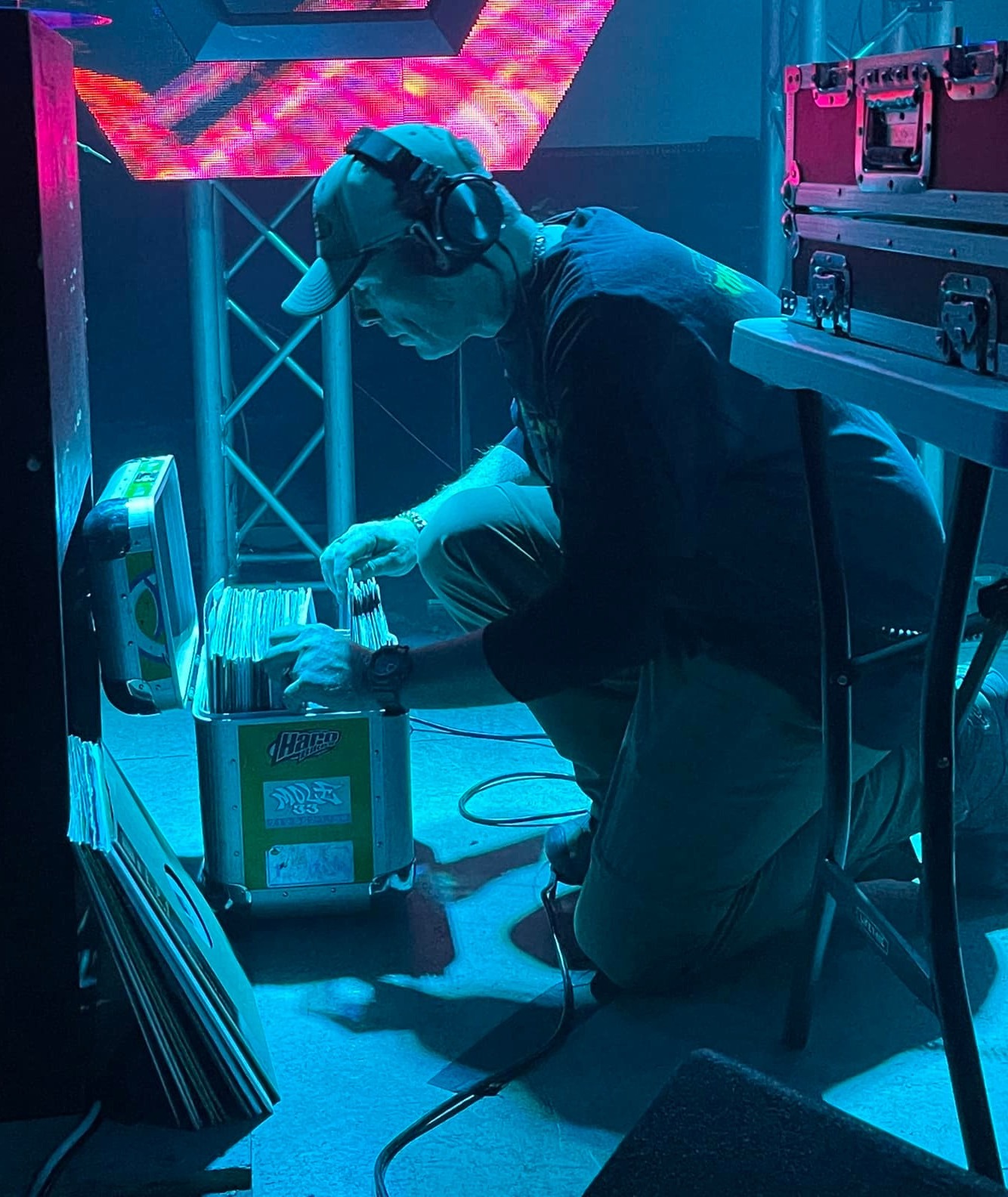 DJ Natural Nate® kneeling and digging through Hario vinyl crate under cyan and pink LED truss lighting